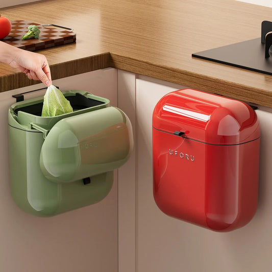 Green wall-mounted trash can with a hand disposing of a plastic bag, next to a red trash can on a kitchen counter.