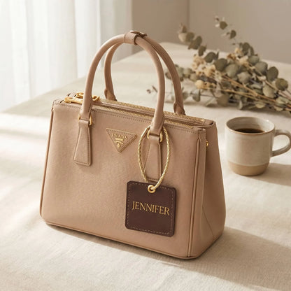 Beige handbag with a Custom name tag on a light surface with a cup and plant in the background