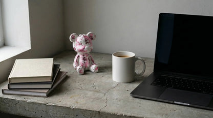 Laptop, mug, books, and teddy bear on a concrete surface near a window.