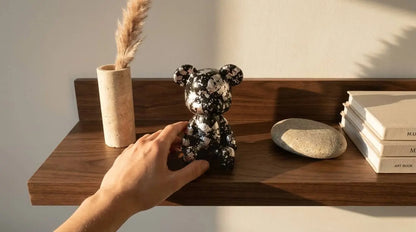 A hand adjusts a Luxe Noir Bear Figurine on a dark wood floating shelf, with a beige vase, books, and a stone nearby in a warm-lit room.